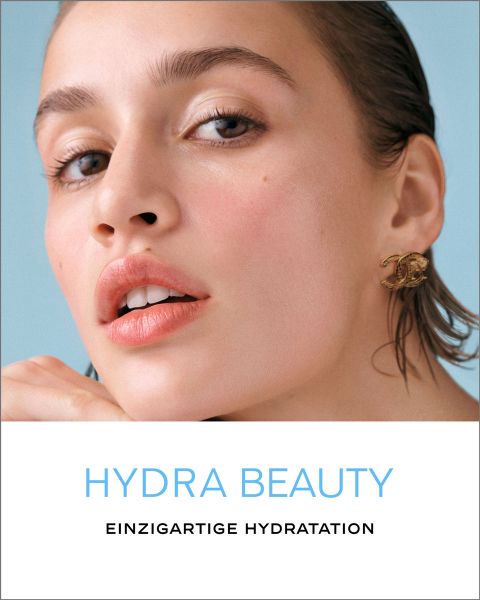 Nahaufnahme einer Frau mit frischer Haut und Nude-Make-up vor hellblauem Hintergrund. Sie trägt einen goldenen Ohrring. Der Text darunter lautet „HYDRA BEAUTY“ und „EINZIGARTIGE HYDRATATION“.
