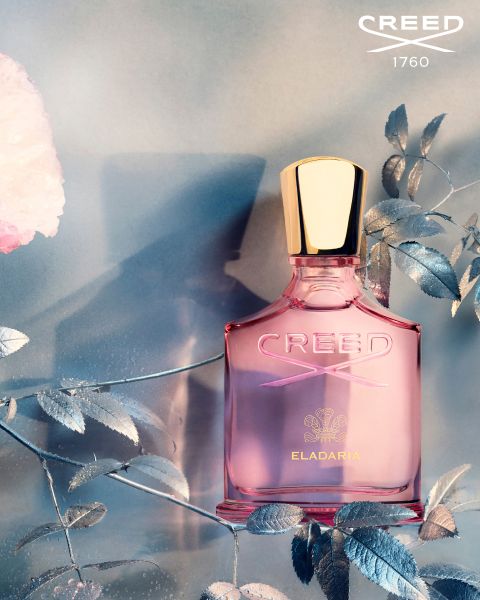 Rosa Parfümflakon mit goldenem Verschluss und der Aufschrift „CREED ELADARIA“ vor einem Hintergrund aus silbernen Blättern und einer zartrosa Blume. Schatten verleihen der eleganten Komposition Tiefe.