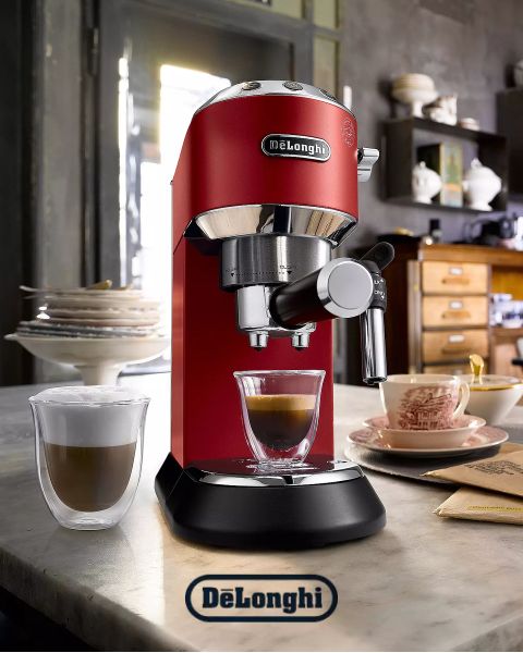 Eine rote De’Longhi-Espressomaschine auf einer Küchentheke gießt Kaffee in eine Glastasse. Im Hintergrund sind eine Tasse Cappuccino, ein Stapel Pfannkuchen und eine gemütliche Küche zu sehen.