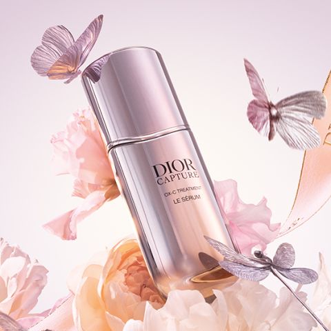 Eine Flasche Dior Capture Totale Le Sérum ist von zartrosa und weißen Blumen und flatternden silbernen und rosa Schmetterlingen umgeben, die vor einem zarten pastellfarbenen Hintergrund stehen.