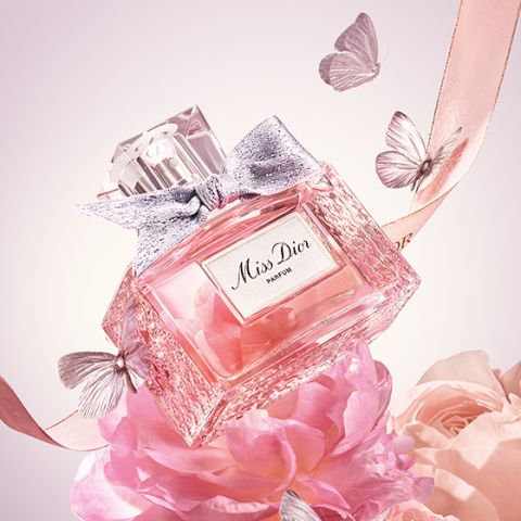 Ein rosafarbener Parfümflakon mit der Aufschrift „Miss Dior“ ist von zarten Schmetterlingen und Bändern umgeben und auf rosafarbenen Blumen platziert. Der Flakon ist mit einer silbernen Schleife verziert. Die Szene vermittelt eine elegante und romantische Atmosphäre.