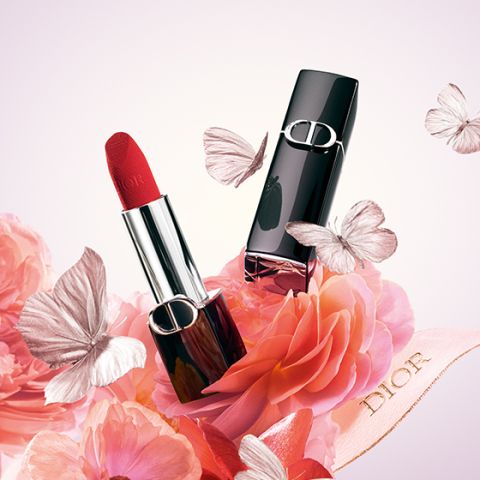 Ein roter Lippenstift in einem silber-schwarzen Etui ist von rosa Blumen und Schmetterlingen umgeben. Der Schriftzug „Dior“ ist auf einem mit den Blumen verschlungenen Band zu sehen. Der Hintergrund ist in einem sanften Farbverlauf aus Rosa und Weiß gehalten.