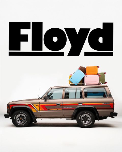 Ein Oldtimer-SUV im Retro-Design mit Gepäck auf dem Dach parkt vor weißem Hintergrund. Über dem Fahrzeug prangt in fetten schwarzen Lettern das Wort „Floyd“.