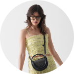 Eine Frau, die ein hellgelbes, ärmelloses Crop-Top mit Rüschen und einen dazu passenden Rock trägt, dazu eine dunkle Sonnenbrille und eine schwarze Umhängetasche, steht vor einem schlichten weißen Hintergrund.