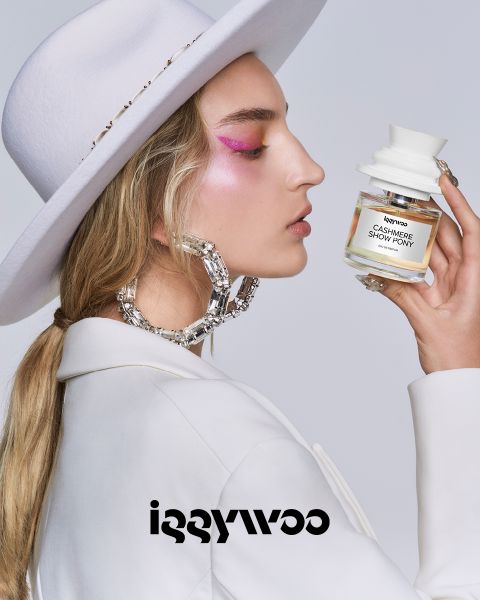Eine Person mit langen Haaren, einem weißen Hut und rosa Augen-Make-up hält eine Flasche mit der Aufschrift „Cashmere Show Pony“ von Iggywoo. Sie trägt große, glänzende Creolen und eine weiße Jacke vor einem schlichten Hintergrund. Unten ist das Iggywoo-Logo zu sehen.