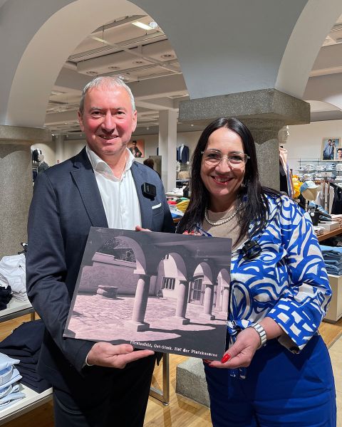 Ein Mann im Anzug und eine Frau in einem blau gemusterten Outfit stehen lächelnd in einem Gebäude und halten ein Buch in den Händen. Das Cover zeigt eine Architekturszene mit Bögen. Im Hintergrund sind Kleiderständer und gewölbte Säulen zu sehen.
