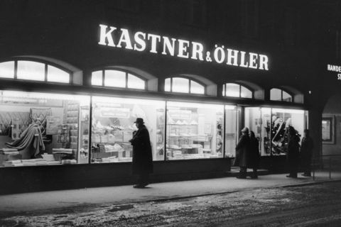 Schwarz-Weiß-Bild eines Kastner & Öhler-Ladens bei Nacht. Vier Personen in langen Mänteln und Hüten stehen auf der verschneiten Straße und betrachten die Schaufenster mit gefalteten Textilien und verschiedenen Artikeln. Der Laden ist hell beleuchtet und bildet einen Kontrast zur dunklen Umgebung.