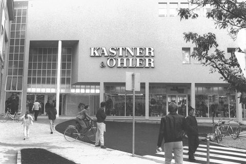 Schwarz-Weiß-Foto von Fußgängern und Radfahrern in der Nähe eines Kastner & Öhler-Geschäfts. Das moderne Gebäude hat große Fenster und eine schlichte Fassade. Im Vordergrund steht ein Baum, rechts ein Laternenpfahl.