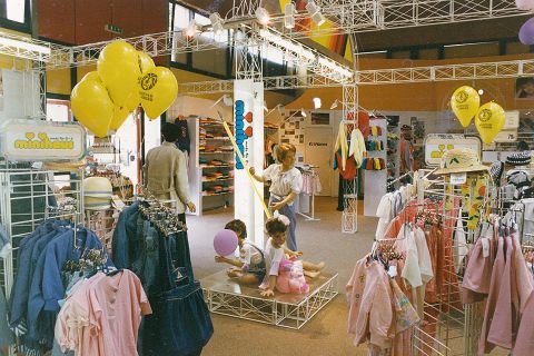 Kinder spielen auf einer Schaubühne in einem farbenfrohen Bekleidungsgeschäft voller Kleiderständer. Leuchtend gelbe Luftballons schweben über den Köpfen, und es werden verschiedene bunte Kleidungsstücke und Freizeitoutfits zum Verkauf angeboten.