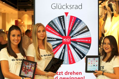 Drei Frauen in weißen G-STAR-Shirts halten Tablets vor einem „Glücksrad“-Display in einem Geschäft. Auf dem Rad sind verschiedene Preise wie Gutscheine und Rabattaktionen zu gewinnen.