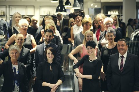 Eine große Gruppe professionell gekleideter Menschen posiert in einem Innenbereich auf einer Rolltreppe. Die Gruppe besteht aus Männern und Frauen mit unterschiedlichen Gesichtsausdrücken. Im Hintergrund ist ein modernes Interieur mit Regalen und Hängelampen zu sehen.