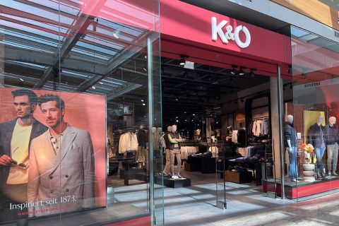 Schaufenster von K&Ö, einem Bekleidungsgeschäft mit Glaseingang. Links im Bild ein großes Poster mit zwei Männern in stylischen Outfits. Im Laden präsentieren Schaufensterpuppen verschiedene Kleidungsstücke. Das Geschäft ist modern eingerichtet und die Kleidung übersichtlich präsentiert.