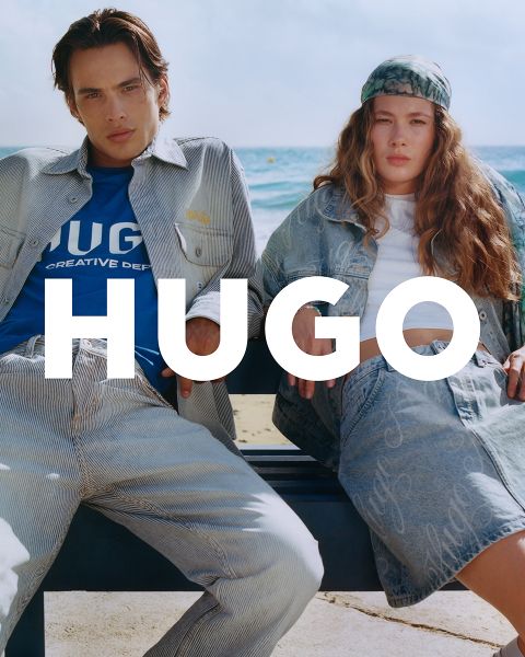 Zwei Models sitzen auf einer Bank am Strand. Beide tragen Jeans-Outfits mit offenen Jacken, unter denen ein blaues T-Shirt und ein weißes Top hervorschauen. In der Mitte prangt deutlich der Schriftzug „HUGO“. Im Hintergrund sind Meer und Himmel zu sehen.