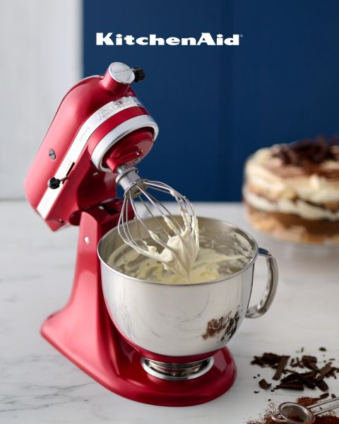 Eine rote KitchenAid-Küchenmaschine mit Schlagsahne auf dem Schneebesen steht auf einer Marmorplatte. Im Hintergrund sind ein geschichtetes Dessert und Schokoladenraspeln zu sehen, darüber steht „KitchenAid“.