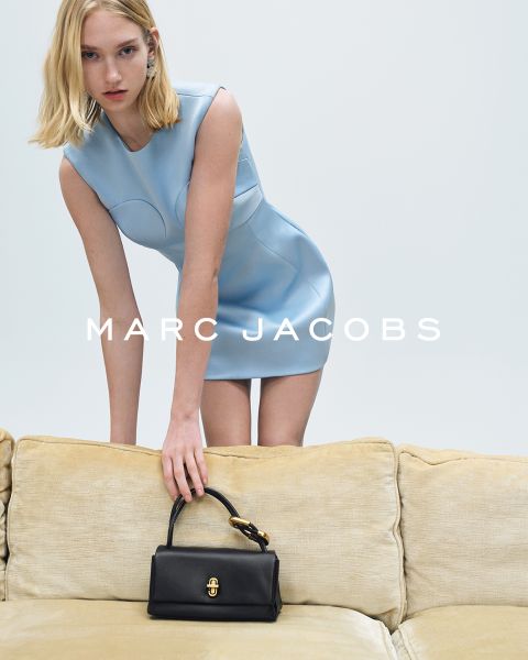 Eine Person mit schulterlangem blondem Haar lehnt sich auf einem beigefarbenen Sofa vor. Sie trägt ein ärmelloses hellblaues Kleid. Sie hält eine schwarze Handtasche in der Hand. Der Schriftzug „MARC JACOBS“ steht in weißer Schrift über dem Bild.