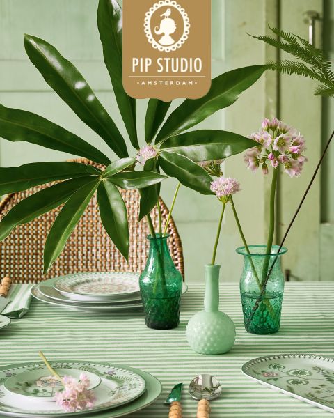 Auf einem Tisch mit grüngestreiftem Stoff stehen Glasvasen mit belaubten Zweigen und rosa Blumen. Teller mit kunstvollen Mustern stehen neben Besteck. Im Hintergrund sind ein Korbstuhl und ein Pip Studio-Logo zu sehen.