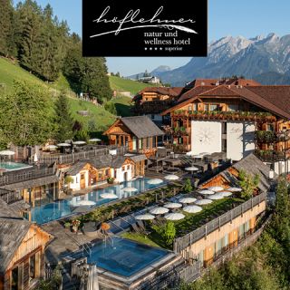 Luftaufnahme des Höflehner Natur- und Wellnesshotels, umgeben von üppig grünen Hügeln und Bergen. Holzgebäude und Terrassen umgeben mehrere Pools mit Sonnenschirmen. Darüber strahlend blauer Himmel. Hotellogo und -name im oberen Bereich.