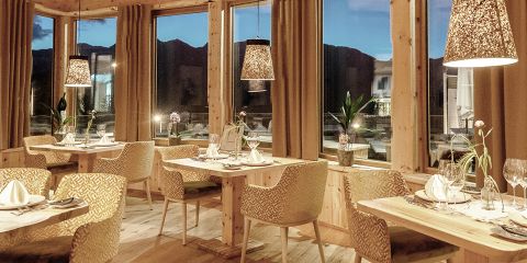Das gemütlich beleuchtete Restaurant mit Holzdekor bietet Tische mit weißen Servietten, Gläsern und Topfpflanzen. Große Fenster geben den Blick in der Abenddämmerung auf Berge und Gebäude frei. Über jedem Tisch hängen Deckenleuchten mit strukturierten Schirmen.
