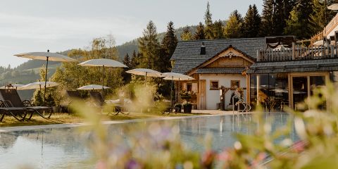 Ein ruhiger Außenpoolbereich mit Sonnenliegen und Sonnenschirmen vor der Kulisse üppiger Bäume und einer rustikalen Holzhütte. Die Szenerie ist in warmes Sonnenlicht getaucht und vermittelt eine friedliche und einladende Atmosphäre.