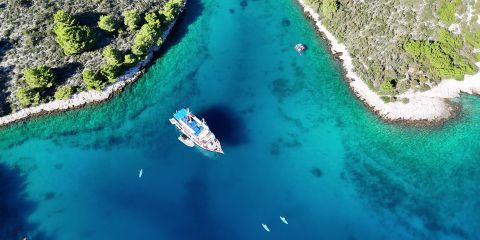 Luftaufnahme einer türkisfarbenen Bucht, umgeben von Grün. In der Mitte liegt ein Segelboot vor Anker, in der Nähe befinden sich kleine Boote. Das klare Wasser gibt den Blick auf den Meeresboden frei, während üppige Bäume das umliegende Land bedecken.