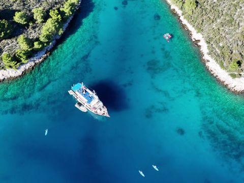 Luftaufnahme einer türkisfarbenen Bucht, umgeben von Grün. In der Mitte liegt ein Segelboot vor Anker, in der Nähe befinden sich kleine Boote. Das klare Wasser gibt den Blick auf den Meeresboden frei, während üppige Bäume das umliegende Land bedecken.