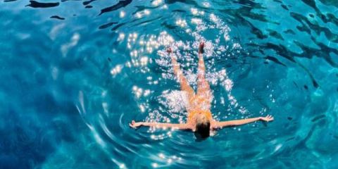 Eine Person schwimmt und treibt auf dem Rücken in klarem, glitzernd blauem Wasser und erzeugt dabei Wellen um sich herum. Sonnenlicht spiegelt sich auf der Wasseroberfläche und erzeugt helle Muster. Die Szene vermittelt ein Gefühl von Entspannung und Ruhe.