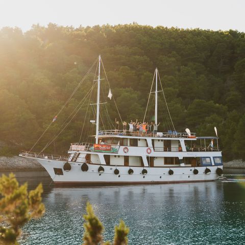 Ein weißes Doppeldeckerboot mit ein paar winkenden Menschen auf dem Oberdeck gleitet über ruhiges Wasser. Umgeben von üppigem Grün und bewaldeten Hügeln wird die Szene von warmem Sonnenlicht von links beleuchtet.