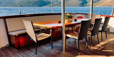 Ein Holztisch mit sechs Stühlen steht auf einem Bootsdeck mit Blick auf ein ruhiges Gewässer. Eine Vase mit Blumen steht auf dem Tisch. Im Hintergrund sind üppige, grüne Hügel unter einem klaren, blauen Himmel zu sehen.