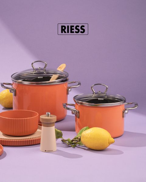 Zwei orangefarbene Kochtöpfe mit Glasdeckeln, ein Holzlöffel und eine Pfeffermühle stehen auf einem violetten Untergrund. Daneben stehen eine Zitrone, grüne Beeren, gestapelte orangefarbene Schalen und beige Teller. Darüber ist der Markenname „RIESS“ zu sehen.