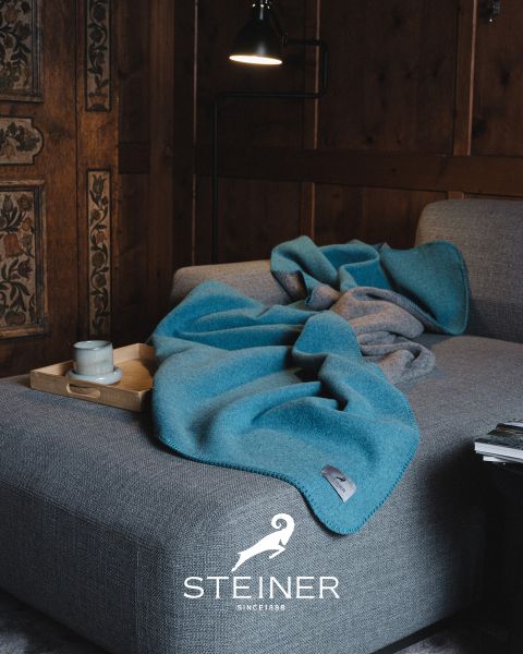 Ein gemütliches Wohnzimmer mit einem grauen Sofa, einer blauen Decke, einem Holztablett mit Tasse und Schüssel, einer Stehlampe und holzgetäfelten Wänden. Unten ist das „STEINER“-Logo zu sehen.
