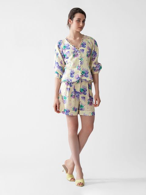 Eine Frau trägt eine hellgelbe Bluse mit Blumenmuster und gewellten Kanten sowie passende Shorts. Sie steht vor einem schlichten weißen Hintergrund, hat ein Bein leicht übereinandergeschlagen und trägt beige Sandalen mit Absatz.