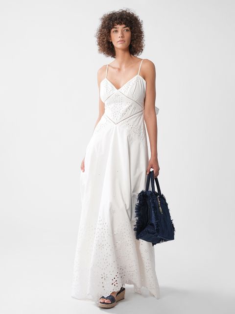Eine Frau mit lockigem Haar trägt ein langes weißes Lochmusterkleid mit dünnen Trägern und hält eine marineblaue strukturierte Handtasche in der Hand. Sie steht vor einem schlichten weißen Hintergrund und trägt Keilsandaletten.