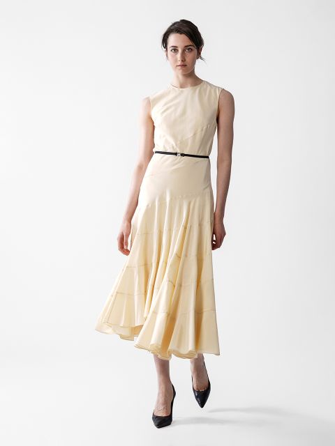 Eine Frau steht vor einem schlichten weißen Hintergrund. Sie trägt ein ärmelloses, hellgelbes Midikleid mit einem schmalen schwarzen Gürtel und schwarzen High Heels. Ihr Gesichtsausdruck ist neutral, und ihr Haar ist locker zurückgebunden.
