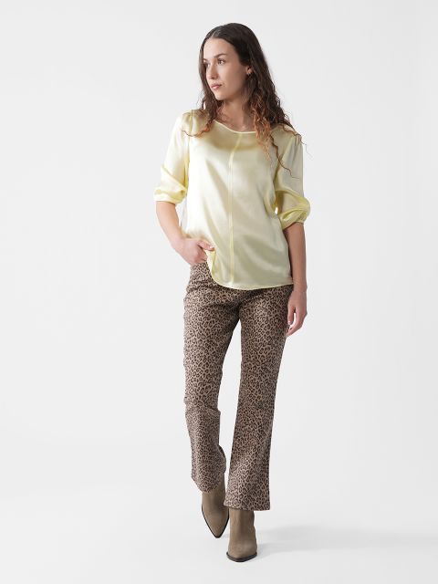 Eine Frau steht vor einem weißen Hintergrund. Sie trägt eine hellgelbe Bluse mit hochgekrempelten Ärmeln, eine Hose mit Leopardenmuster und taupefarbene Stiefeletten. Sie hat langes, welliges braunes Haar und blickt zur Seite.