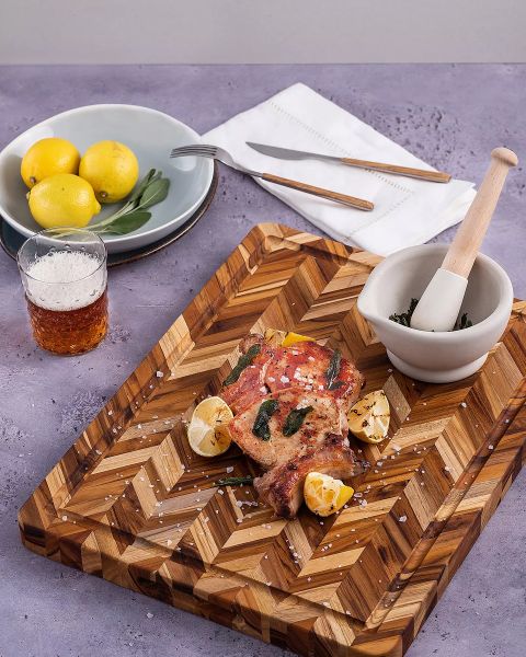 Ein hölzernes Schneidebrett mit gewürztem, gekochtem Fleisch, Zitronenspalten und grobem Salz. Daneben stehen ein Mörser mit Kräutern, ein Glas mit Getränk, ein Teller mit Zitronen sowie Gabel und Messer auf einer Serviette, alles auf einer violetten Oberfläche.