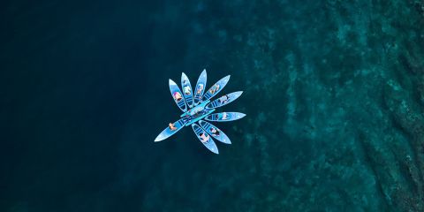 Luftaufnahme von sieben Kajaks mit Menschen darin, die auf klarem, seichtem Wasser ein kreisförmiges Muster bilden. Die Kajaks sind blau und bilden einen auffälligen Kontrast zum aquamarinblauen Hintergrund.