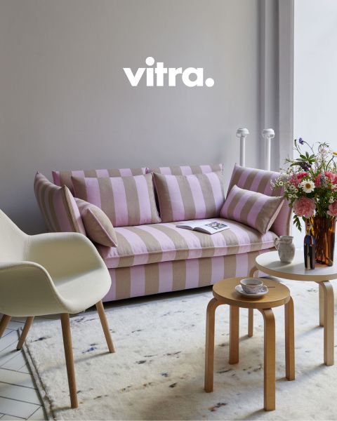 Ein modernes Wohnzimmer verfügt über ein rosa-beige gestreiftes Sofa, einen weißen Stuhl mit Holzbeinen und einen runden Couchtisch aus Holz. Auf dem Tisch steht eine Vase mit bunten Blumen, darüber steht „vitra.“ an der Wand. Der Boden ist mit einem weichen Teppich bedeckt.