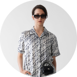 Eine Person trägt ein kurzärmeliges, schwarz-weiß gemustertes Hemd, eine schwarze Sonnenbrille und eine kleine schwarze Handtasche vor einem schlichten weißen Hintergrund.