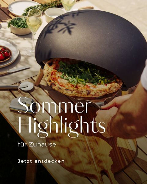 Eine Person nimmt eine frisch gebackene Pizza aus einem modernen Outdoor-Pizzaofen auf einen Holztisch, der mit Tellern, Weingläsern und frischen Zutaten gedeckt ist. Der Text lautet: „Sommer-Highlights für Zuhause. Jetzt entdecken.“.