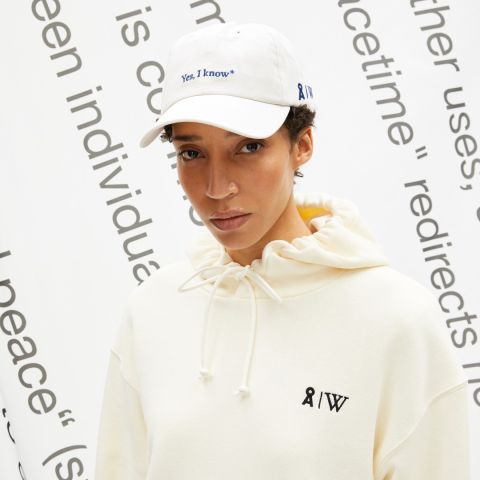Eine Person trägt einen weißen Kapuzenpullover mit Kordelzug und eine weiße Kappe mit der Aufschrift „Ja, ich weiß*“ und steht vor einem weißen Hintergrund mit teilweise sichtbarem schwarzen Text.