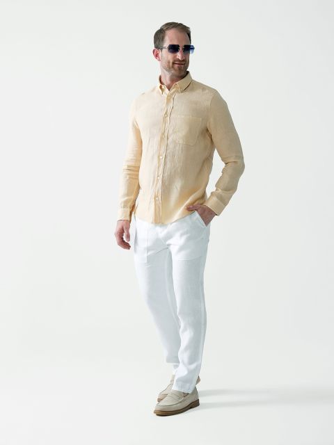 Ein Mann mit Sonnenbrille, hellgelbem Langarmhemd, weißer Hose und beigen Slippern steht vor einem schlichten weißen Hintergrund, blickt zur Seite und lächelt.