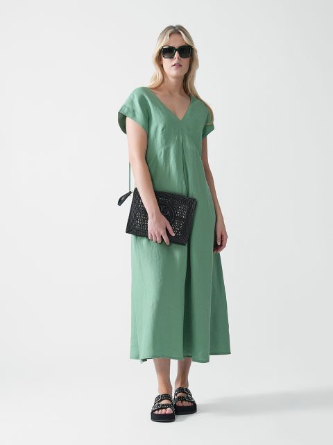 Eine Frau in einem lockeren, hellgrünen Kleid mit V-Ausschnitt, schwarzen Sandalen und einer dunklen Sonnenbrille hält eine schwarze Clutch mit Struktur und steht vor einem schlichten weißen Hintergrund.