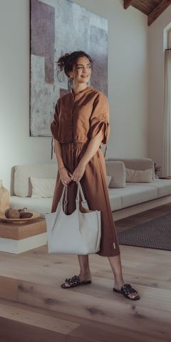 Eine Frau in einem braunen Kleid und schwarzen Sandalen steht drinnen auf einem Holzboden und hält eine große weiße Einkaufstasche in der Hand. Sie lächelt und blickt zur Seite. Im Hintergrund sind ein modernes Sofa und Kunstwerke zu sehen.