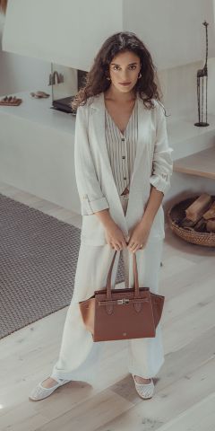Eine Frau mit langen Locken steht in einem hellen Anzug und einem gestreiften Hemd im Haus. Sie hält eine braune Handtasche in der Hand und blickt in die Kamera. Der Raum hat helle Holzböden und ist minimalistisch eingerichtet.
