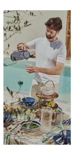An einem sonnigen Tag gießt ein Mann in einem weißen Hemd Wasser aus einem blauen Krug in ein Glas an einem mit blauem und weißem Geschirr, Trauben und Salat gedeckten Tisch im Freien neben einem Swimmingpool.