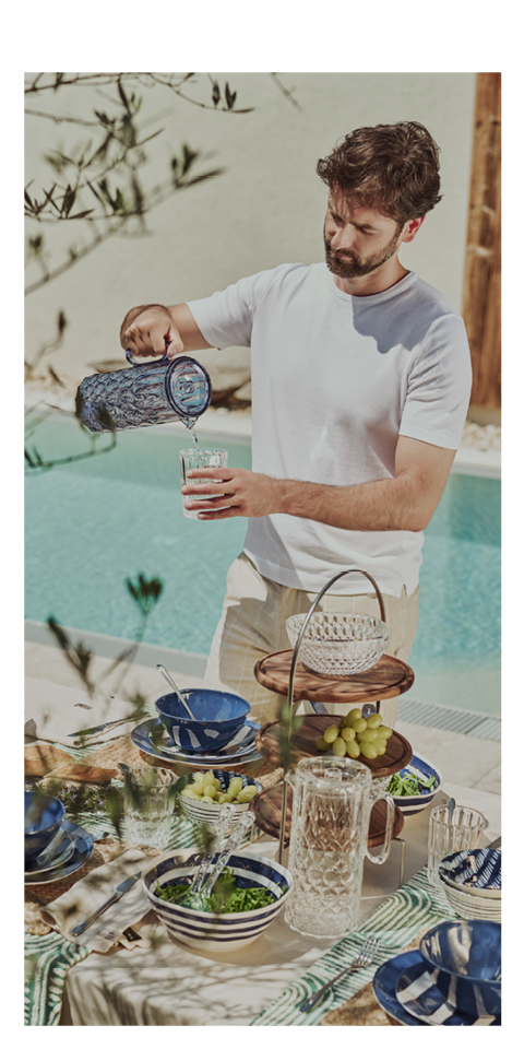 An einem sonnigen Tag gießt ein Mann in einem weißen Hemd Wasser aus einem blauen Krug in ein Glas an einem mit blauem und weißem Geschirr, Trauben und Salat gedeckten Tisch im Freien neben einem Swimmingpool.