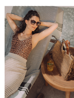Eine Frau mit Sonnenbrille und Badeanzug mit Leopardenmuster liegt im Freien auf einem gestreiften Stuhl. In der Nähe steht auf einem hölzernen Beistelltisch ein gehäkeltes Überkleid, eine gewebte Tasche und ein Glas mit einem Getränk.