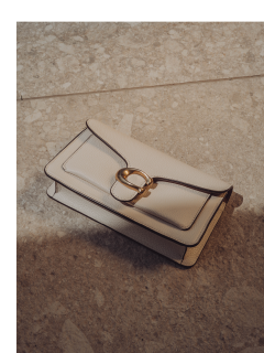 Eine beige Clutch aus Leder mit einer goldenen Schnalle liegt auf einer hellen, teilweise im Schatten liegenden Oberfläche aus Steinfliesen.