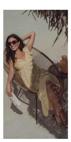Eine Frau in gelbem Kleid und Sonnenbrille entspannt sich in einem geflochtenen Stuhl im Freien und lächelt in die Sonne. Sie hält eine weiße Clutch in der Hand, neben ihr auf einem kleinen Tisch ein Getränk und ein Krug.