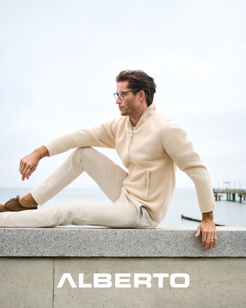 Ein Mann mit Brille, einem beigen Pullover mit Reißverschluss und einer hellen Hose sitzt auf einem Steinvorsprung am Meer. Er blickt zur Seite, unter ihm steht in weißer Schrift "ALBERTO". Der Hintergrund ist wolkenverhangen und ein Pier ist zu sehen.